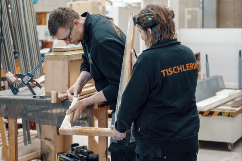 Tichlerin hält einen Holzrahmen an dem ein Tischler arbeitet