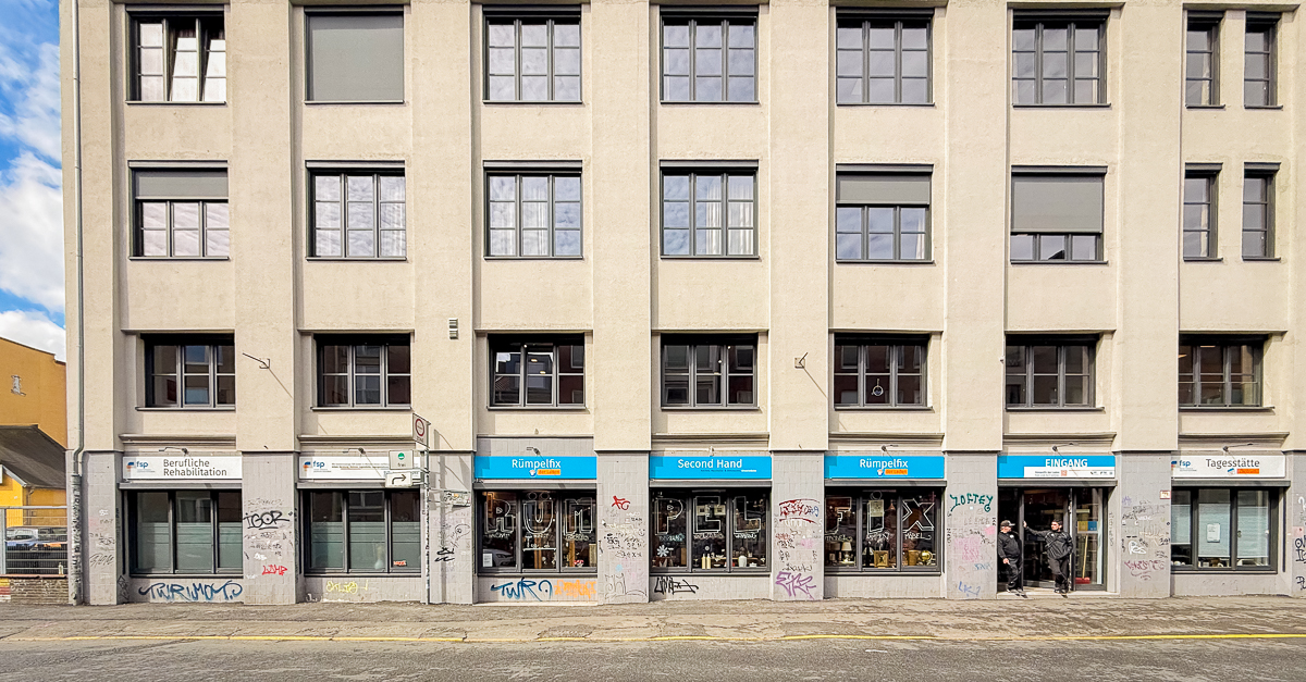 Foto des Gebäude der Bremer Straße 42. Das Foto zeigt viele Fenster der drei Stockwerke. Zwei Personen stehen im Erdgeschoss vor dem Eingang von Rümpelfix der Laden, und reden.